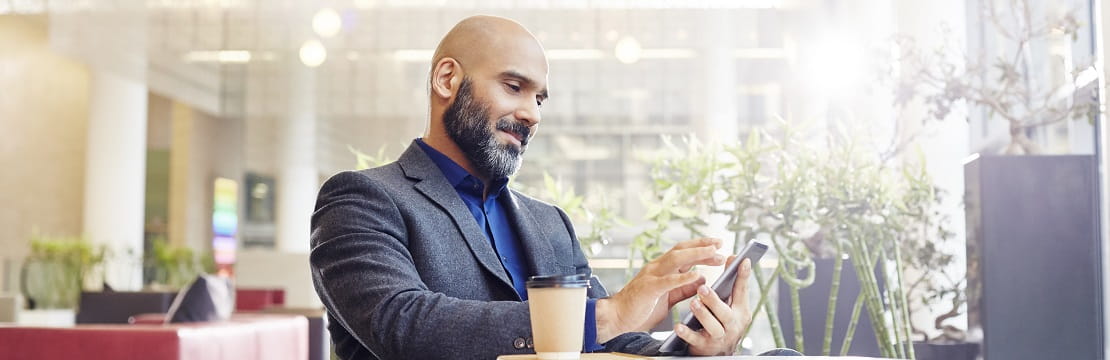 A businessman scrolls through his phone. He is seated at a small table with a cup of coffee beside him.